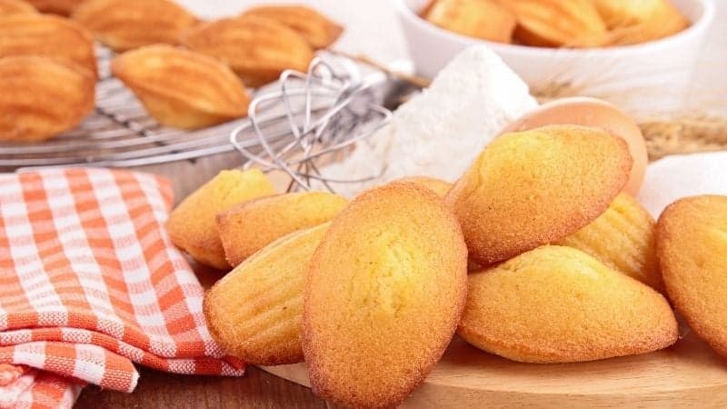 Des madeleines vegan qui sortent du four posées sur une table en bois avec une serviette à carreaux rouges et blancs